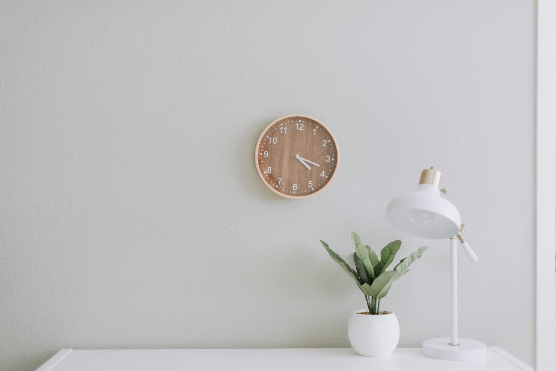 Minimalist interior with a wooden wall clock, white desk lamp, and small green plant on a clean white table against a light wall