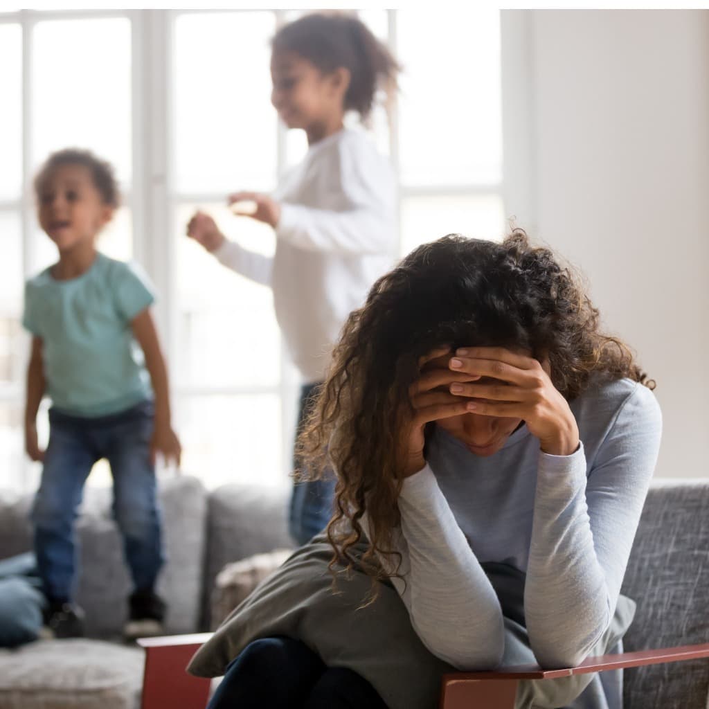 Mother sitting with head in hands appearing stressed while children play in the background, representing parenting challenges addressed in family therapy.